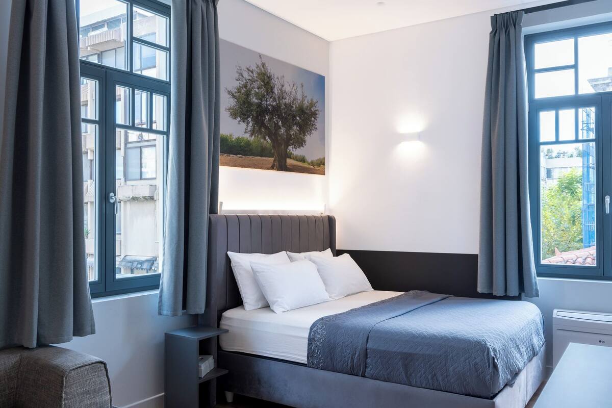 A modern bedroom with a large bed, grey curtains, a painting of a tree, and a view of buildings through the window.