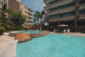 A swimming pool with a hot tub, surrounded by lounge chairs and palm trees, with a multi-story building in the background.