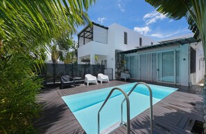 A modern outdoor swimming pool area with a wooden deck, lounge chairs, and a white building in the background. - New & Cozy Loft with Pool in Cap Malheureux (Cap Malheureux)
