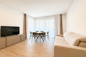 A modern living room with a flat-screen TV, a sofa, a dining table with chairs, and sheer curtains. - Aurora Sand 3 - Carraro Immobiliare Jesolo (Lido di Jesolo)