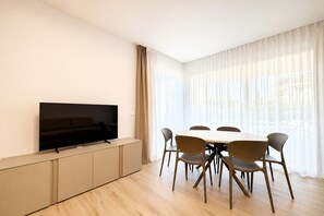 Ein modernes Wohnzimmer mit einem Flachbildfernseher auf einer Kommode, einem Esstisch mit Stühlen und großen Fenstern mit durchsichtigen Vorhängen.