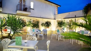 An outdoor patio area with white chairs and tables, surrounded by greenery and a building with arches and a balcony.