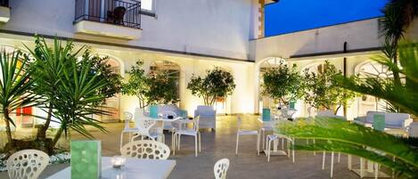 An outdoor patio area with white chairs and tables, surrounded by greenery and a building with arches and a balcony.