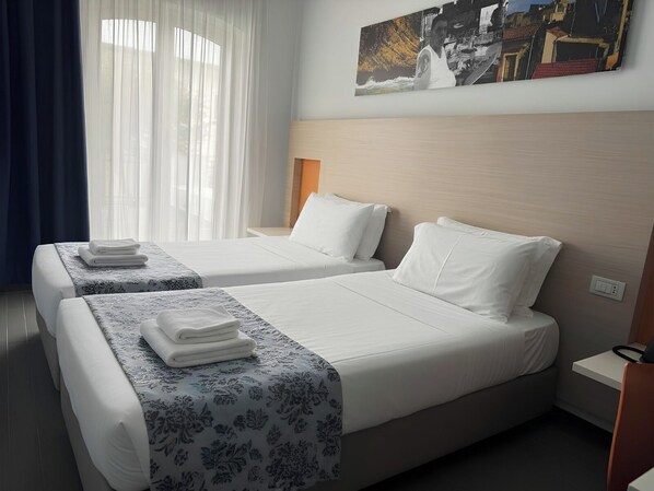 A hotel room with two beds, a large window with sheer curtains, a painting on the wall, and a bedside table with a lamp. - Hotel Kaos (Villaggio Pirandello)