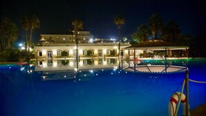 A nighttime view of a hotel with a swimming pool, a covered area with seating, and a building with multiple windows.