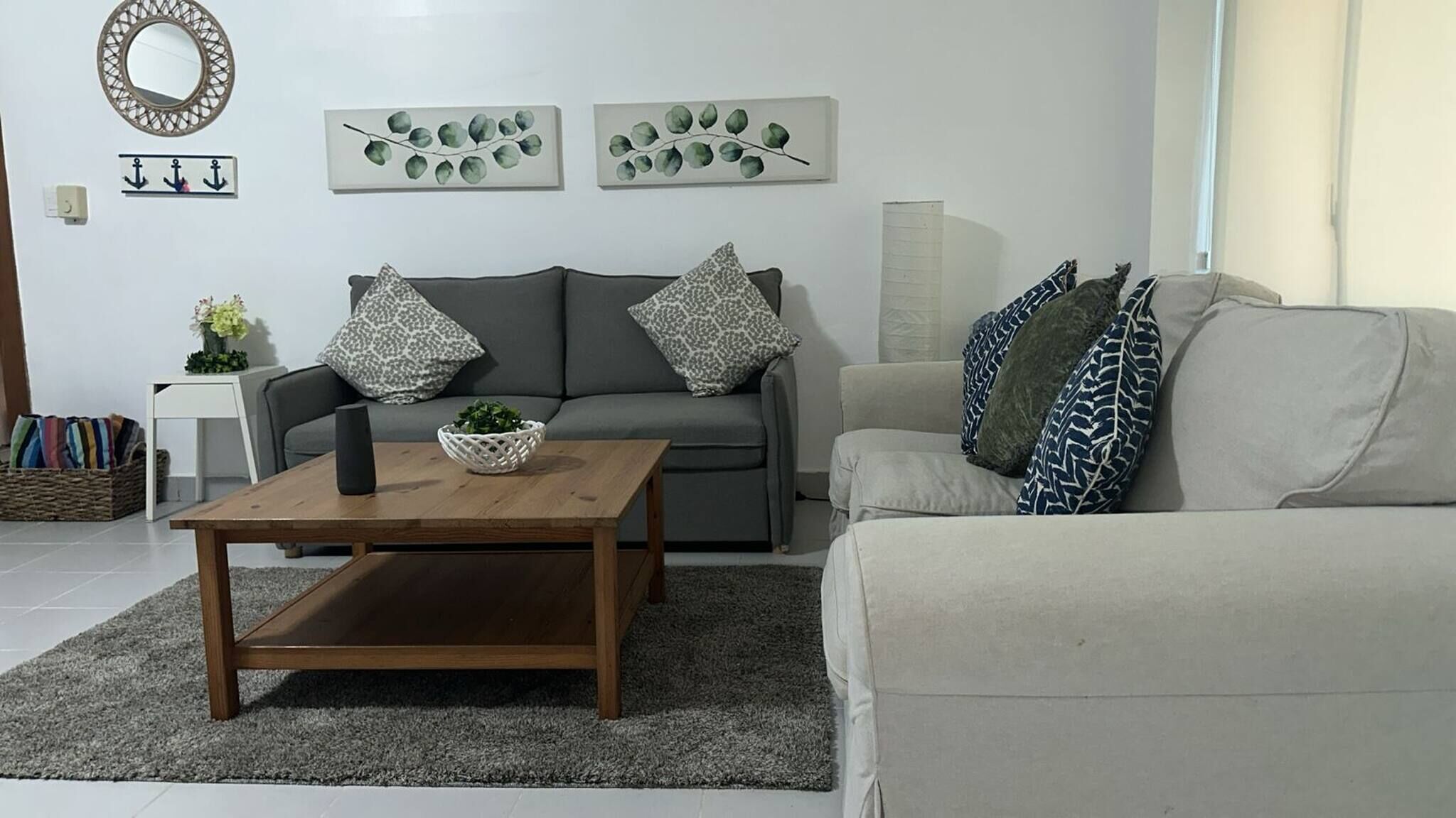 A modern living room with a grey sofa, a wooden coffee table, and a grey rug. There are two wall-mounted artworks depicting eucalyptus leaves and a decorative mirror.