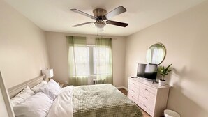Ein Schlafzimmer mit Bett, Kommode, Fernseher, Deckenventilator und einem Fenster mit Vorhängen.