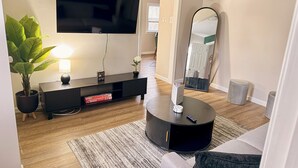 Ein modernes Wohnzimmer mit Flachbildfernseher, einer Konsolentisch, einem runden Beistelltisch mit Fernbedienung, einer Topfpflanze und einem Spiegel.