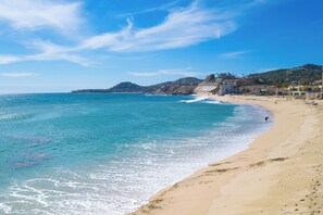 A beach with clear blue water, sandy shore, and buildings in the background. - Beachfront Paradise Mykonos 204A (San José del Cabo)