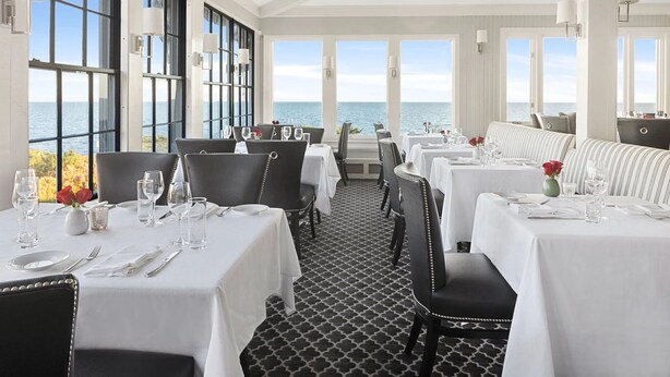 Ein Restaurant mit gedeckten Tischen, großen Fenstern mit Meerblick und einem gemusterten Boden.