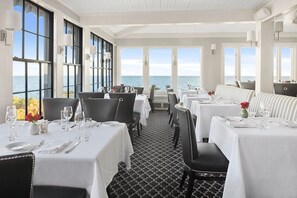 En restaurang med bord dukade för middag, stora fönster med utsikt över havet och ett mönstrat golv.