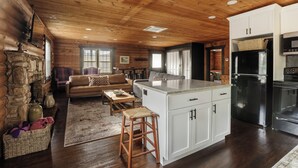 Interior acogedor de una cabaña con techo de madera, chimenea de piedra, zona de estar con sofá y mesa de centro, y cocina con armarios blancos y electrodomésticos de acero inoxidable.