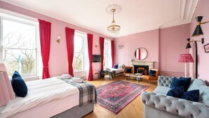 Un dormitorio de color rosa con cama, sofá, chimenea y un amplio ventanal con cortinas.