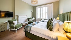 A hotel room with a large bed, a green armchair, a small round table, a television, and a window with blinds. - Triple Room with Private Bathroom (2nd Floor - Room 7) (Edinburgh)