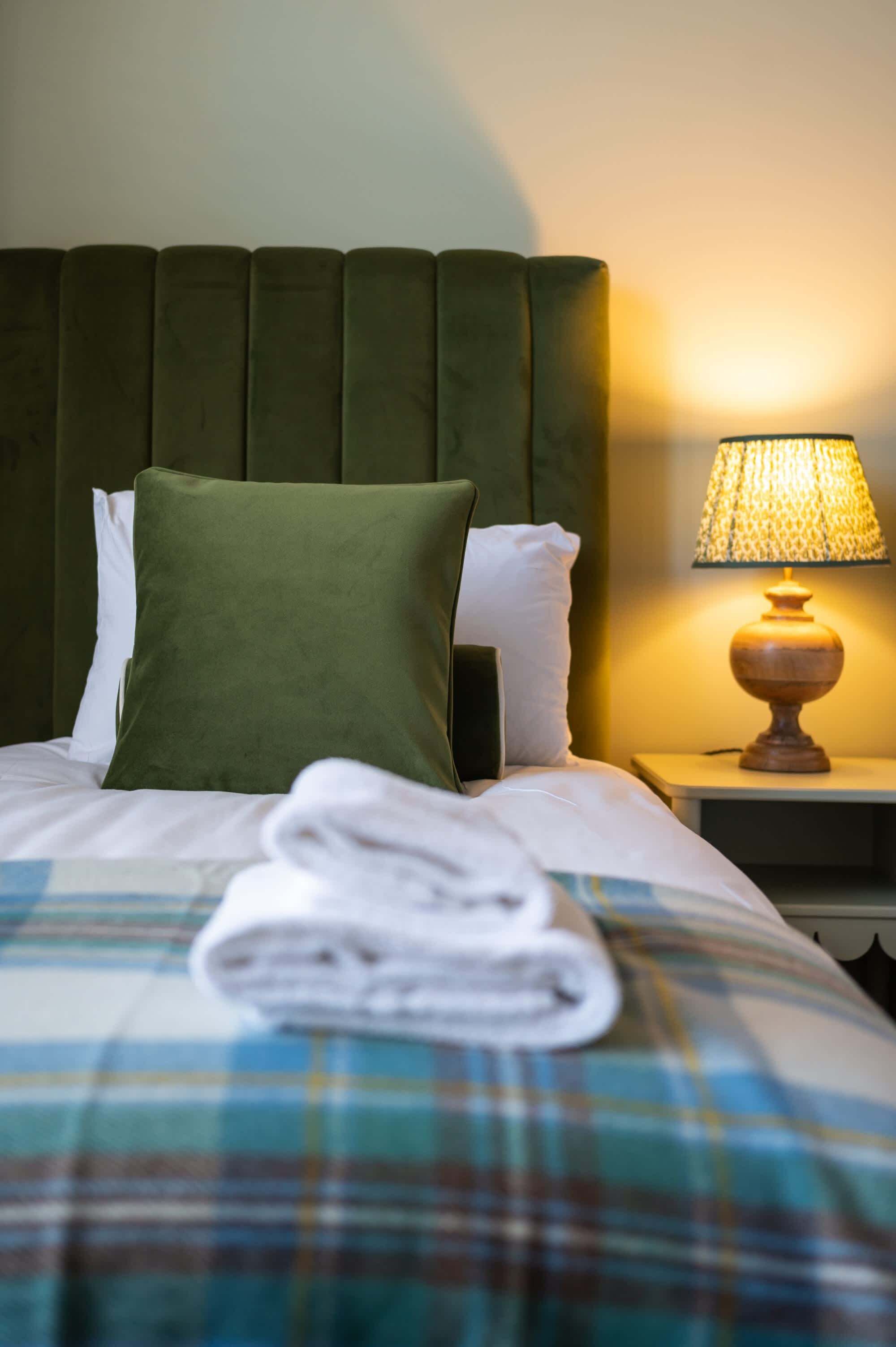 Una cama bien hecha con una manta escocesa, una almohada verde y una toalla doblada sobre la cama. Una lámpara está sobre una mesita de noche al lado de la cama.