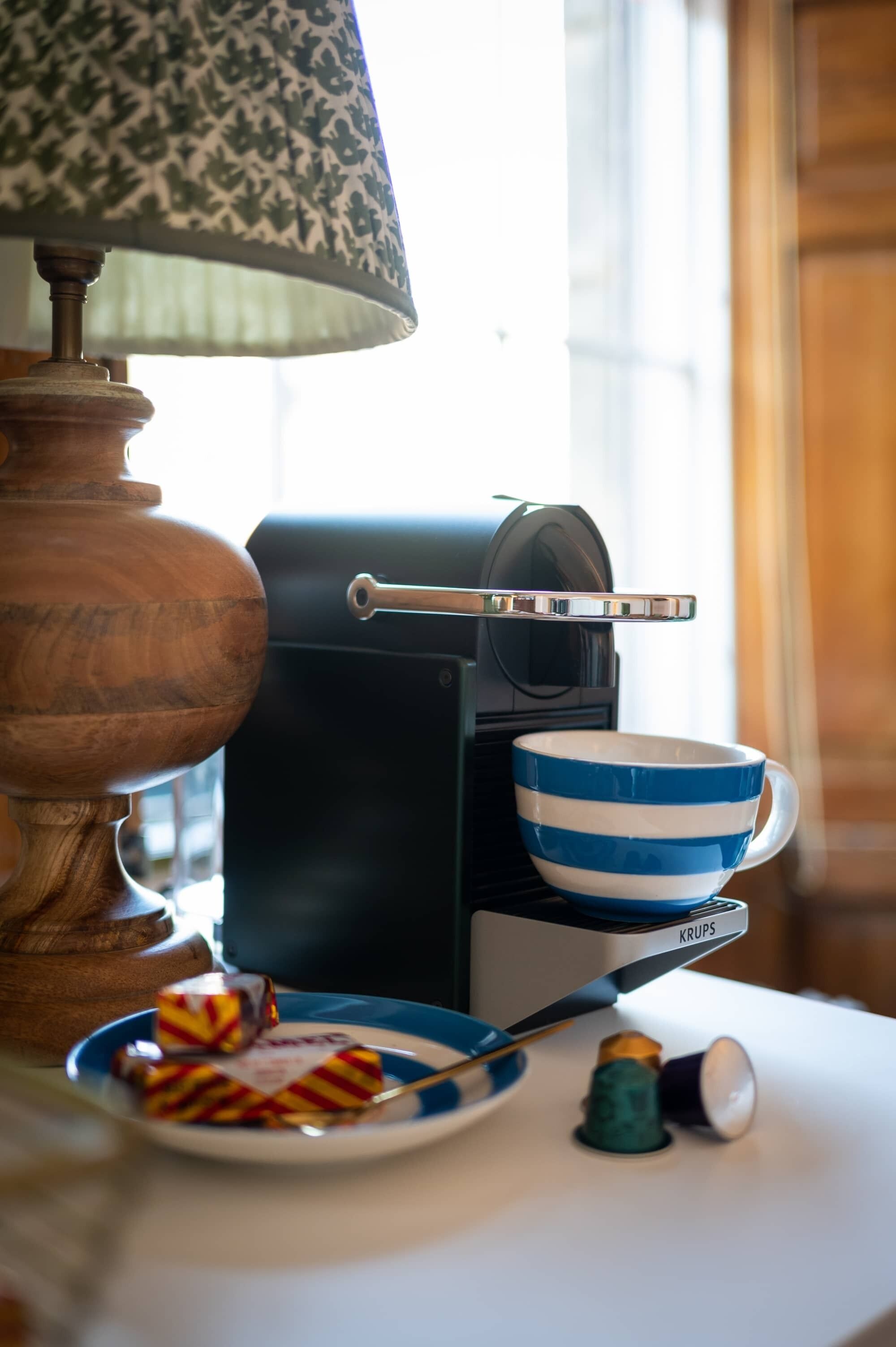 A coffee maker with a blue and white striped mug on its drip tray, a plate with striped candies, and a lamp with a patterned shade.