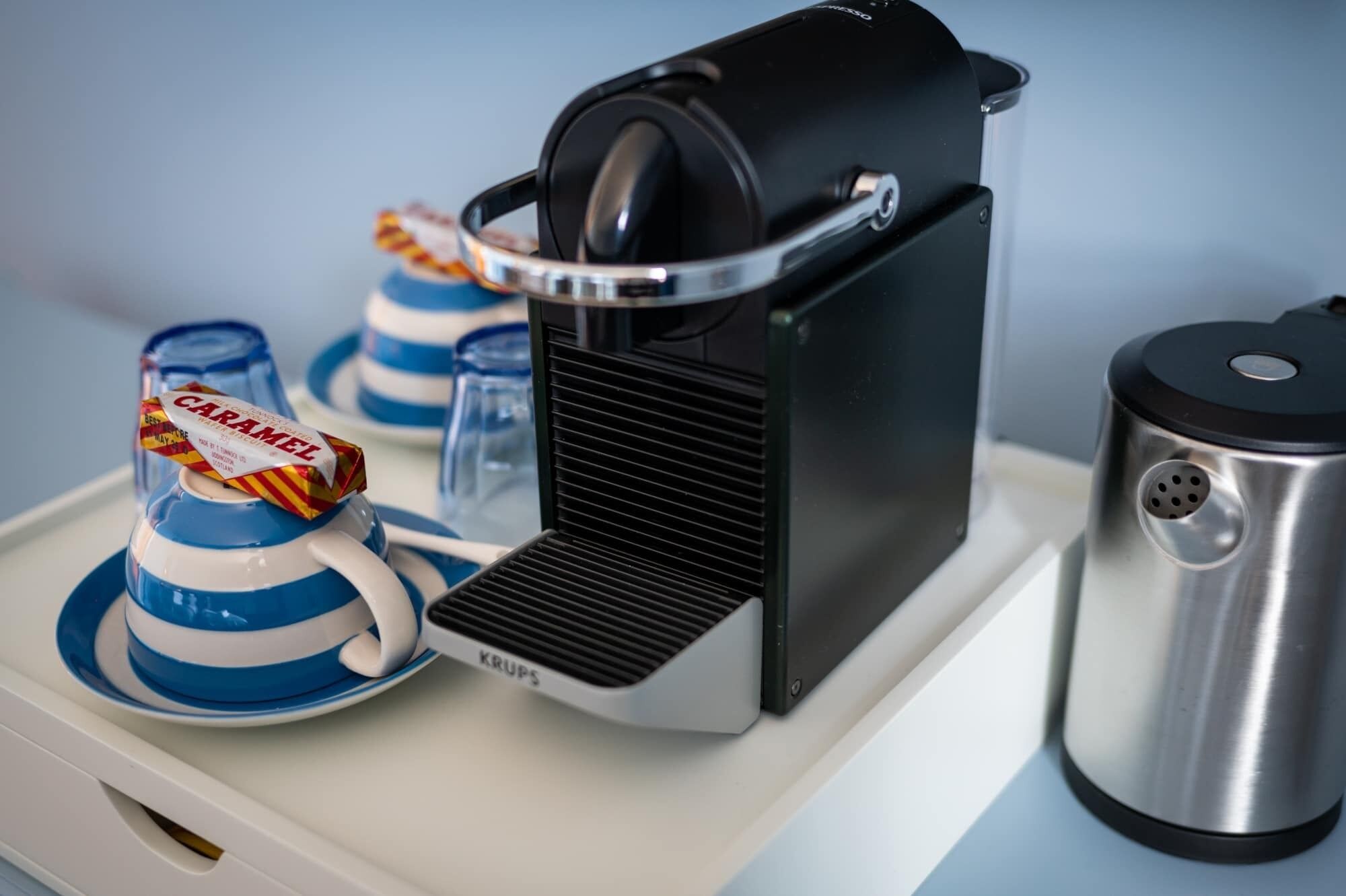 En kaffemaskin med en svart och silverfärgad design, en behållare för vatten i rostfritt stål och två blå koppar med en röd etikett.
