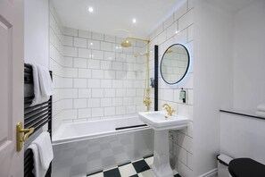 Een moderne badkamer met een witte betegelde muur, een zwart-wit geruite vloer, een witte badkuip, een gouden douchekop, een wit wastafelmeubel en een zwart toilet.