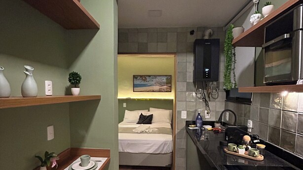 Eine kompakte Küche mit Mikrowelle, Herd und Regalen mit Dekoration. Im Hintergrund ist ein Bett zu sehen, und die Wand ist grün.