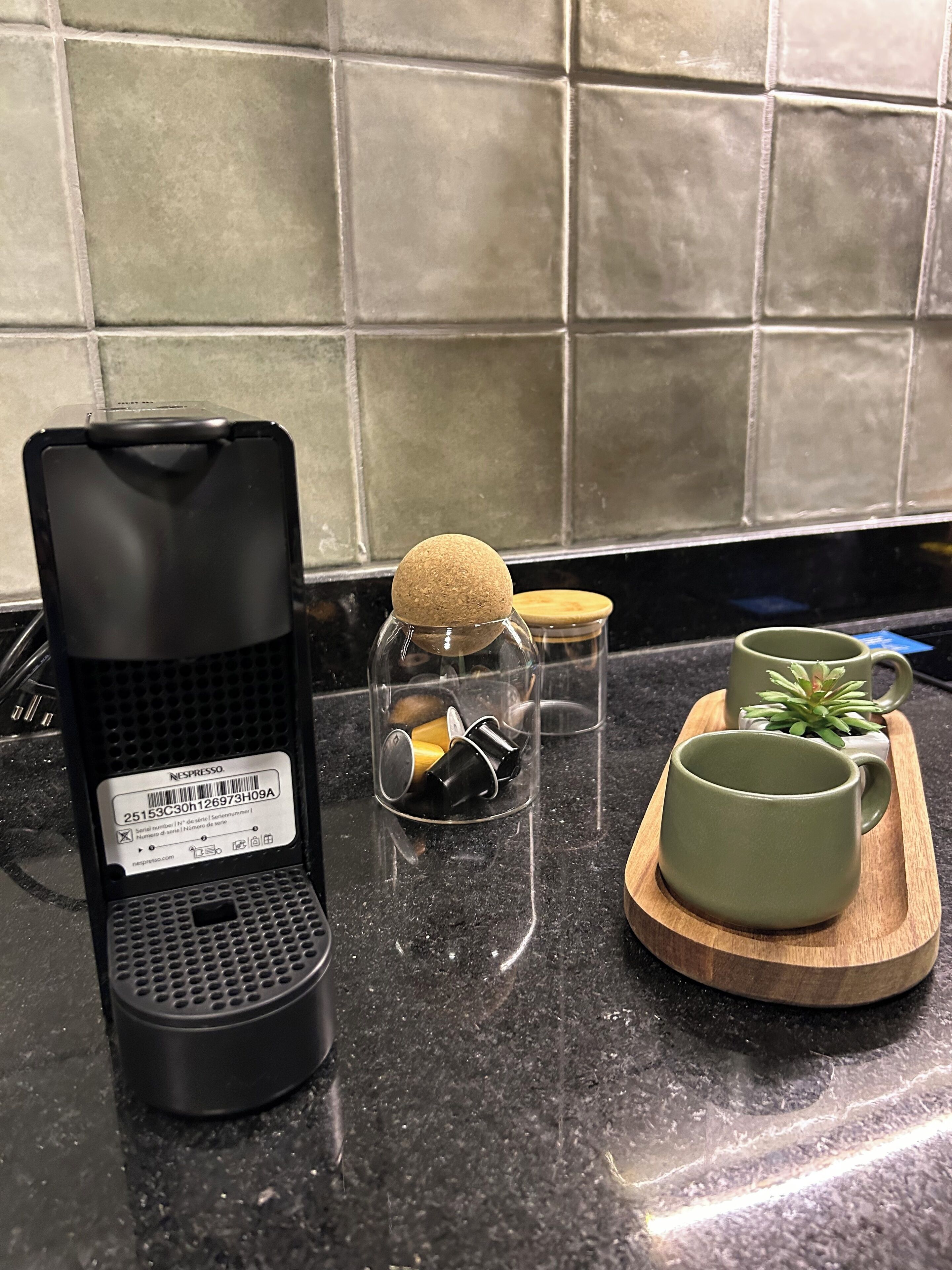 Eine Küchenarbeitsplatte mit einer Kaffeemaschine, einem Glasbehälter mit Korkverschluss und zwei grünen Tassen auf einem Holztablett.