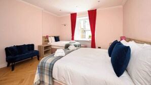 A hotel room with two beds, a sofa, a window with red curtains, and a wooden floor. - Deluxe Family Room (Room 3 - Lower Ground Floor) (Edinburgh)