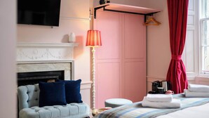 Quarto de hotel com sofá acolchoado, abajur, cama com toalhas dobradas e guarda-roupa rosa.