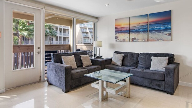 Ein Wohnzimmer mit einem Glastisch, zwei grauen Sofas und einem großen Gemälde, das einen Sonnenuntergang über dem Meer zeigt.