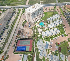 An aerial view of a hotel complex with multiple buildings, a swimming pool, tennis courts, and surrounding greenery.