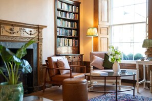 Uma biblioteca acolhedora com poltronas de couro, lareira, estante de livros e uma mesa com livros e um vaso de flores.