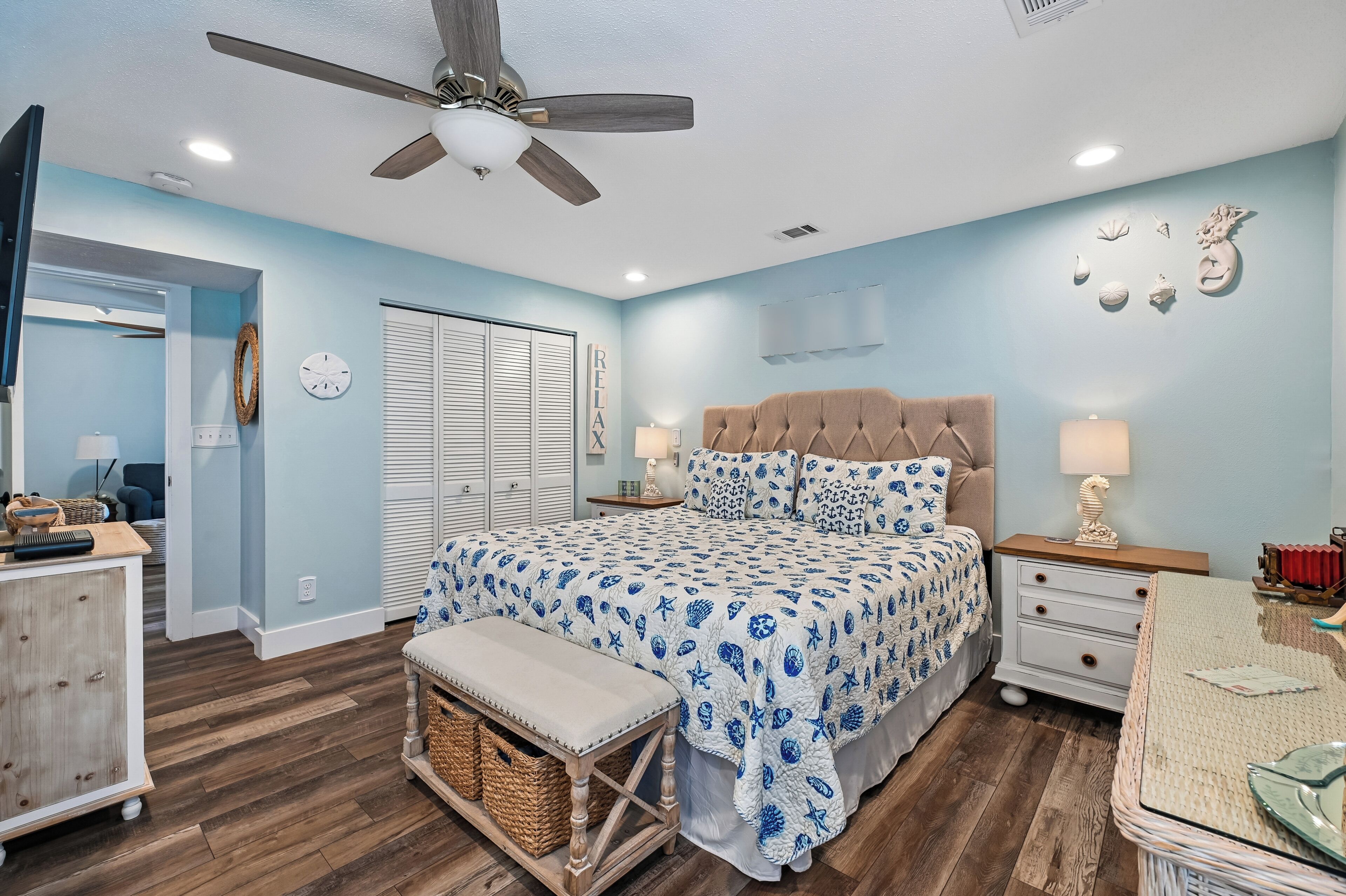 A bedroom with a bed, a bench, a nightstand, a dresser, a ceiling fan, and a door.