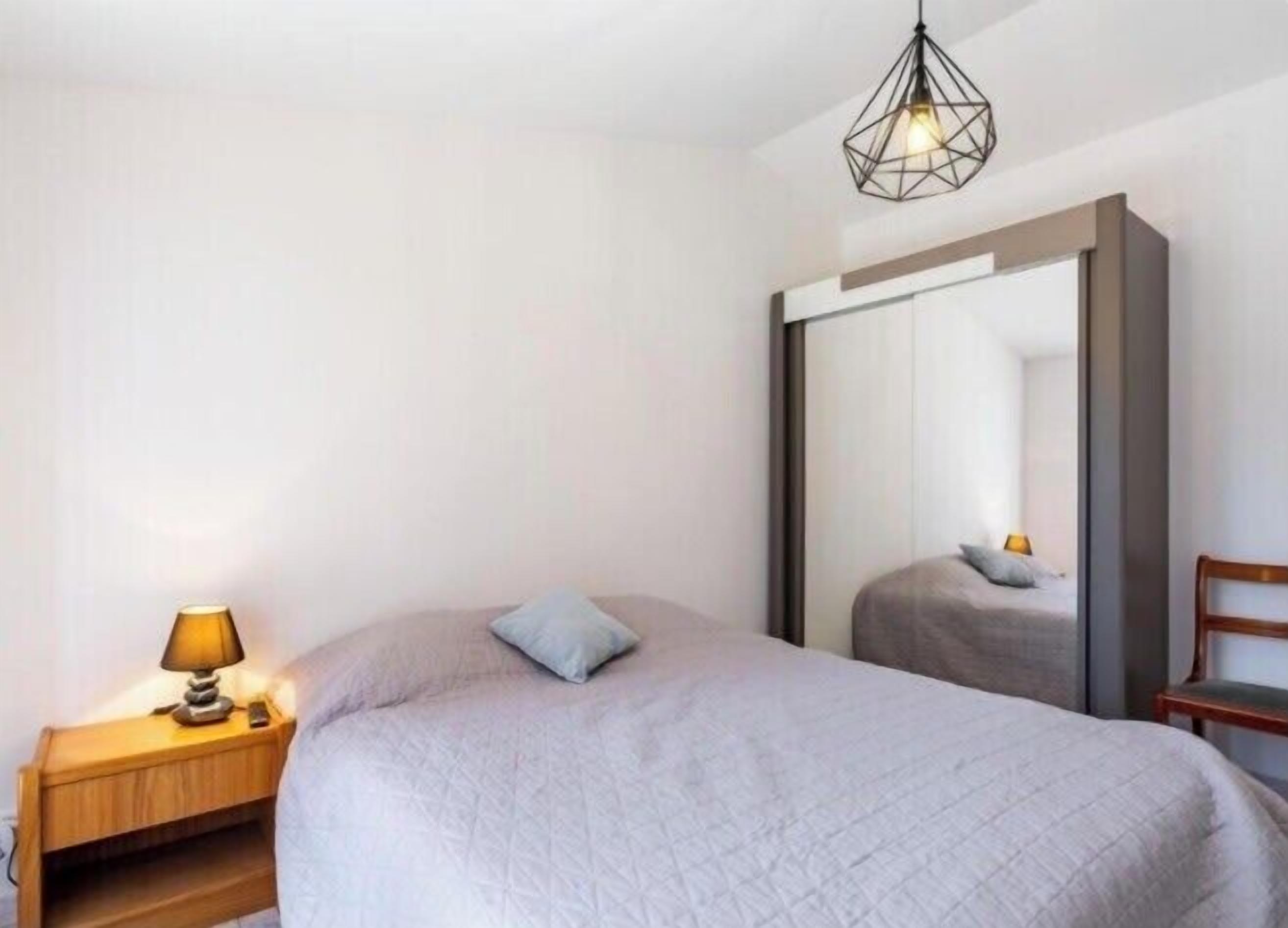 A bedroom with a bed, a nightstand with a lamp, a large mirror, and a hanging light fixture.