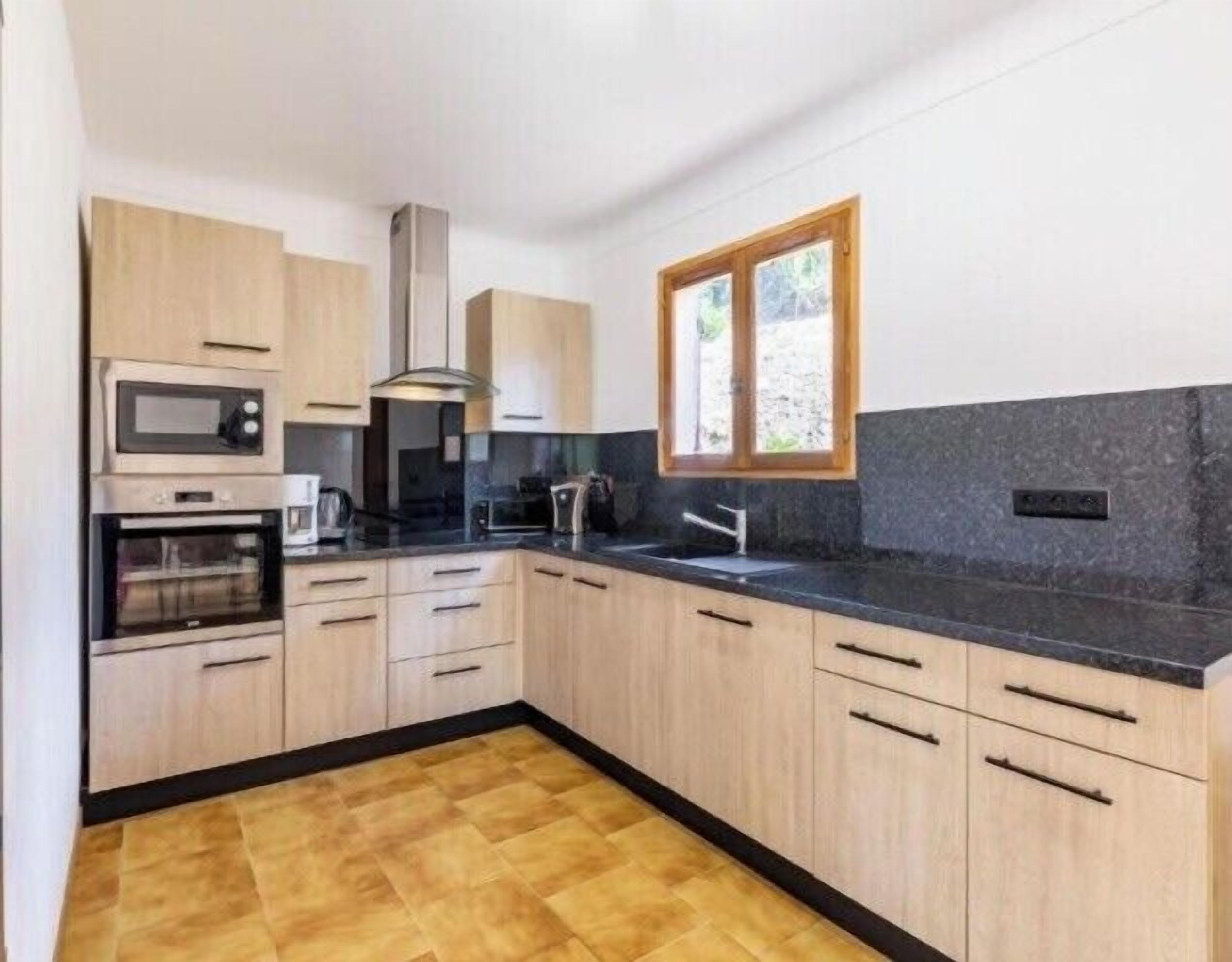 A modern kitchen with wooden cabinets, a black countertop, a built-in oven, a microwave, a window, and a sink.