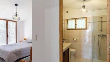 Une chambre à coucher comprenant un lit, une chaise et un luminaire suspendu. La salle de bain est équipée d’une douche, de toilettes et d’un lavabo.