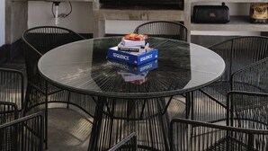 Eine Terrassengruppe im Freien mit einem runden Glastisch, umgeben von schwarzen Stuhlgestellen aus Draht, einem eingebauten Grill und einem JBL-Lautsprecher.