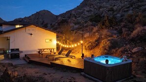 A cozy outdoor patio with a hot tub, lounge chairs, and string lights against a mountainous backdrop. - High Point · 6-Acres, Sweeping Views, Hot Tub (Joshua Tree)