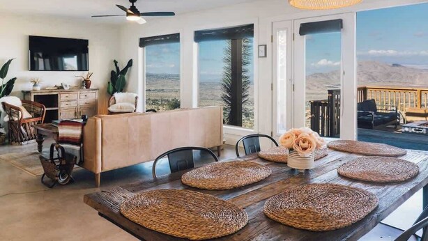 A spacious dining area with a wooden table, woven placemats, and a view of the desert landscape through large windows. - High Point · 6-Acres, Sweeping Views, Hot Tub (Joshua Tree)