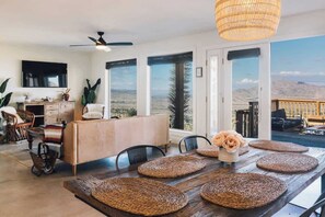 A spacious dining area with a wooden table, woven placemats, and a view of the desert landscape through large windows. - High Point · 6-Acres, Sweeping Views, Hot Tub (Joshua Tree)