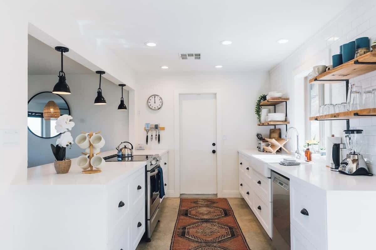 A modern kitchen with white cabinetry, pendant lighting, a clock, and a kitchen island with a built-in oven and dishwasher.