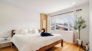 A bedroom with a wooden bed, white bedding, a swan decoration, a nightstand, a potted plant, and a window with a view of buildings. - 2-bedroom condo in beautiful Montréal I Bright, Modern & Cozy (Montréal)