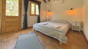 A bedroom with a wooden door, a large window with curtains, a bed with patterned bedding, two bedside tables, and an air conditioning unit. - Detached Single - Storey Stone house in Datça Karaincir - Karaincir Houses 1 (Datça)