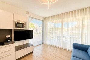 Ein modernes Wohnzimmer mit Fernseher, Mikrowelle und Kaffeemaschine auf einer weißen Kommode. Ein blaues Sofa und ein großes Fenster mit durchsichtigen Vorhängen sind ebenfalls vorhanden.