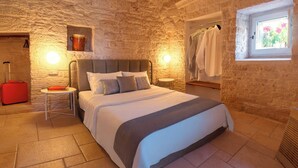Une chambre confortable avec un mur en pierre, un lit doté d’une tête de lit grise, une valise rouge et une fenêtre ornée de fleurs.