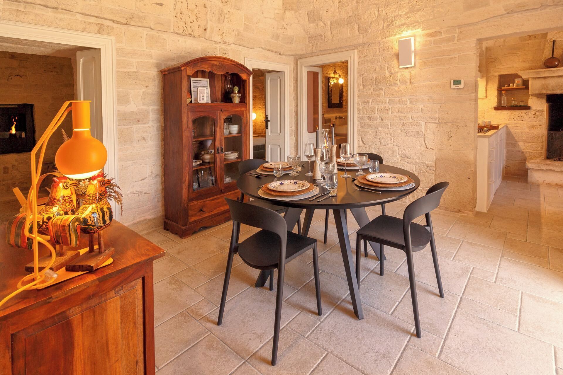 A dining area with a round table set for four, surrounded by black chairs. A wooden cabinet with glass doors is visible in the background.