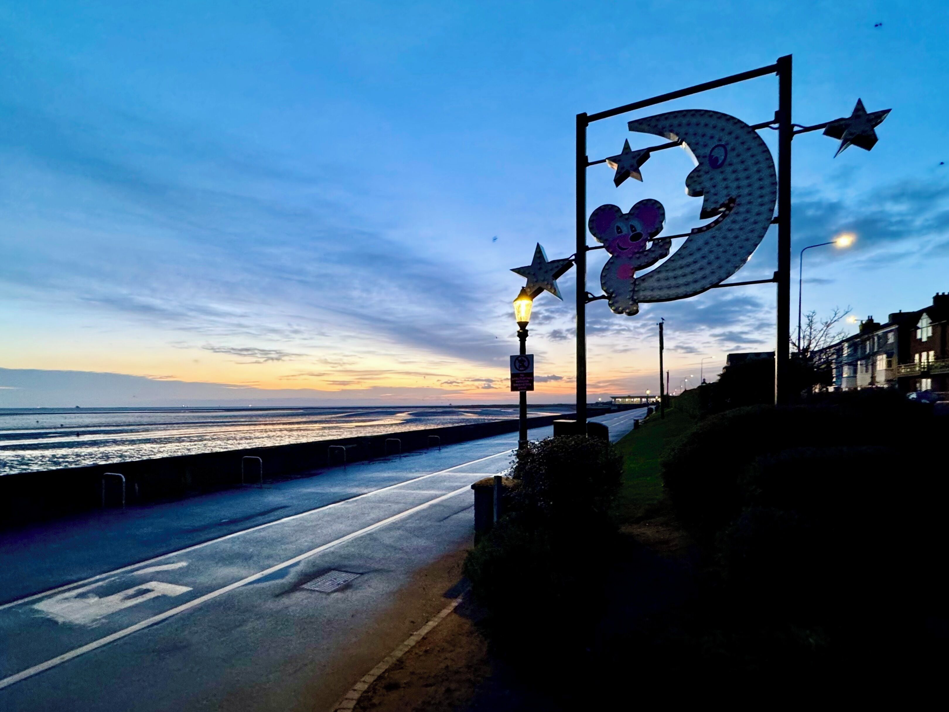 A coastal road with a decorative sign featuring a moon and stars, a street lamp, and a view of the sea at sunset.