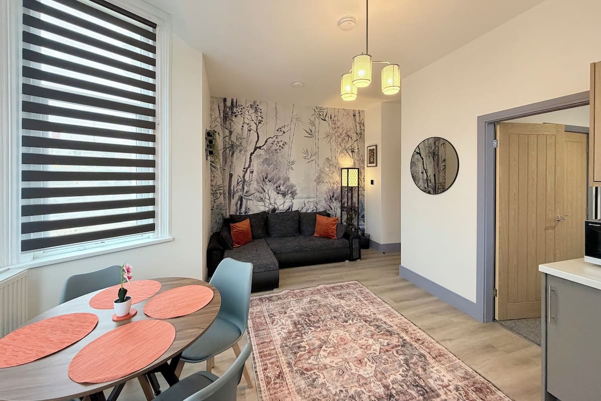 A modern living room with a sofa, a dining table with chairs, a patterned rug, and a wall with a bamboo mural.
