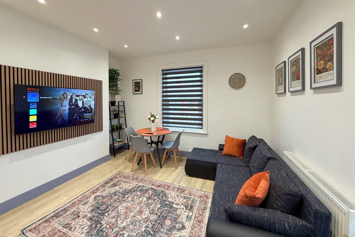 A modern living room with a flat-screen TV, a dining area with a table and chairs, a sofa with orange cushions, and a patterned rug.
