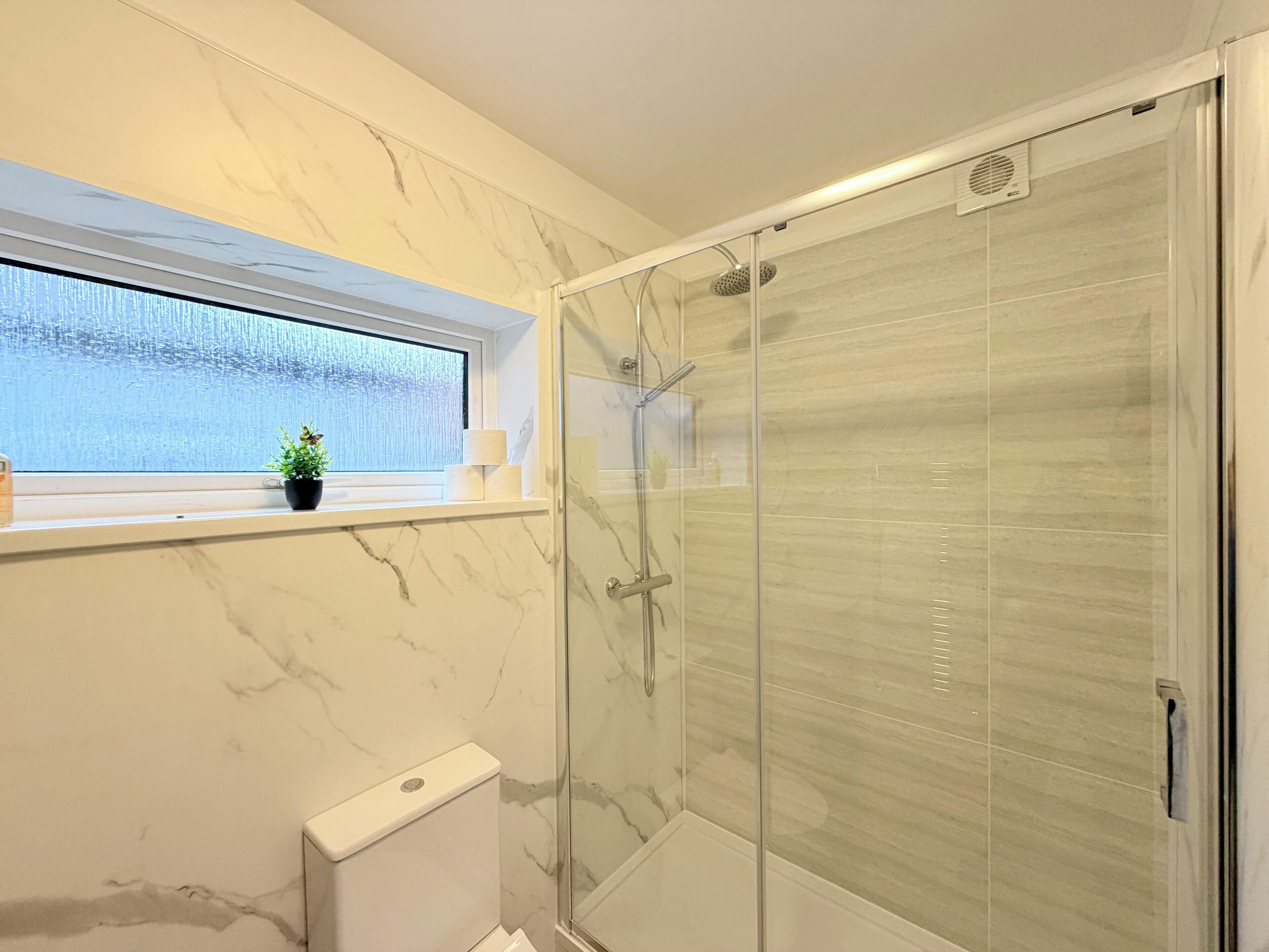 Une salle de bain avec une douche à parois de verre, des toilettes, une fenêtre avec une plante et des murs en marbre.