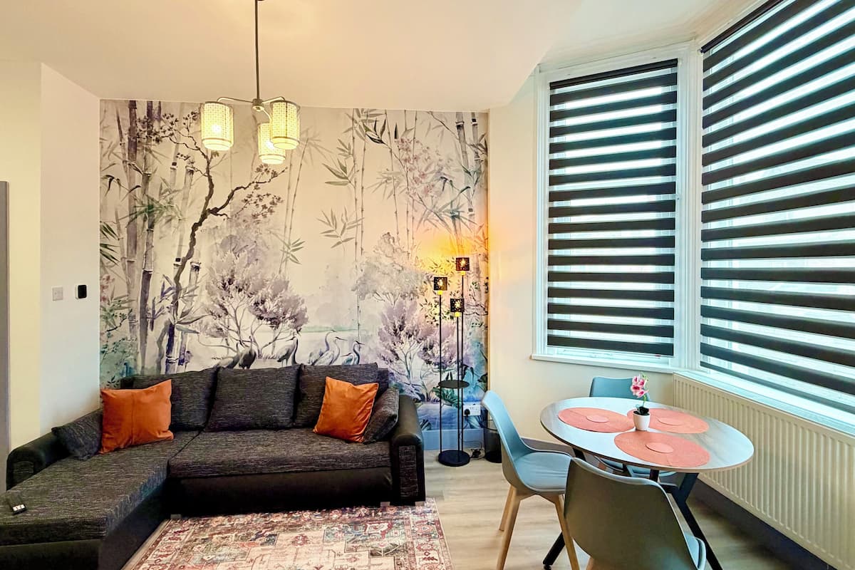 A living room with a sofa, a dining table with chairs, a patterned rug, a wall with a nature-themed mural, and a window with blinds.