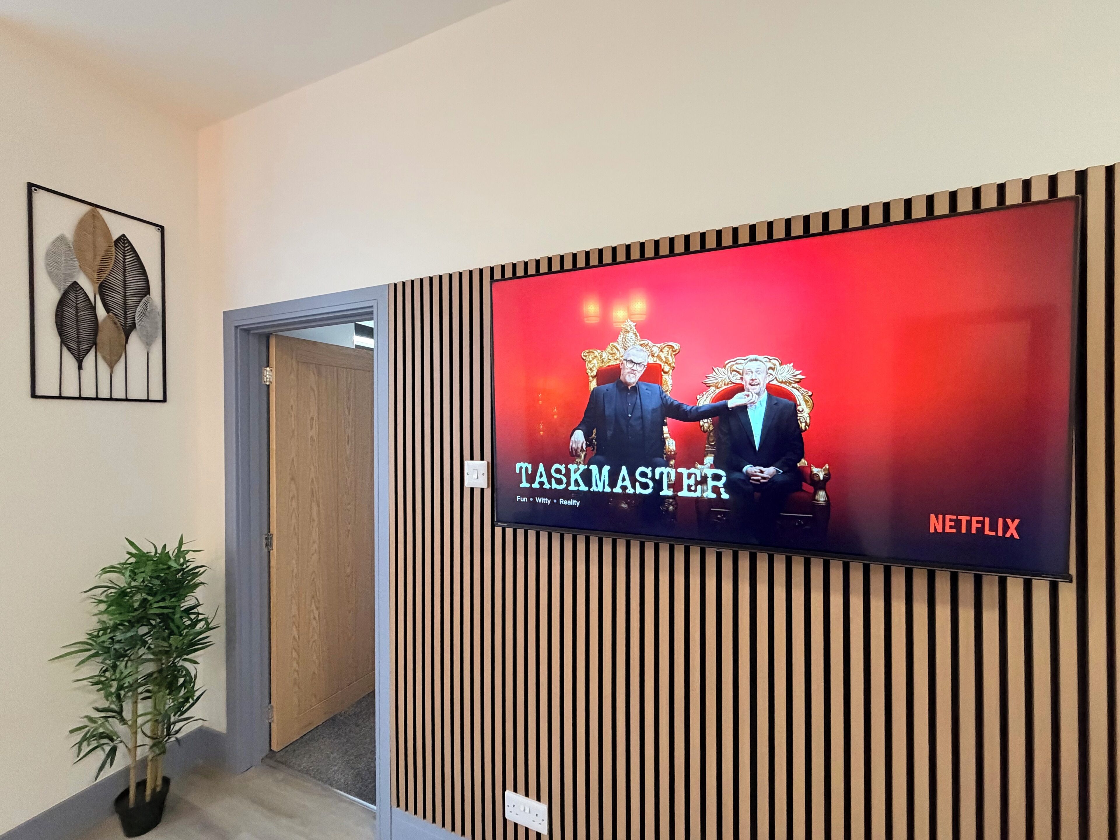 A television displaying 'TASKMASTER' with a Netflix logo, mounted on a wall with vertical wooden slats.