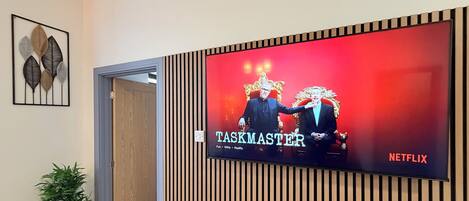 A television displaying 'TASKMASTER' with a Netflix logo, mounted on a wall with vertical wooden slats.